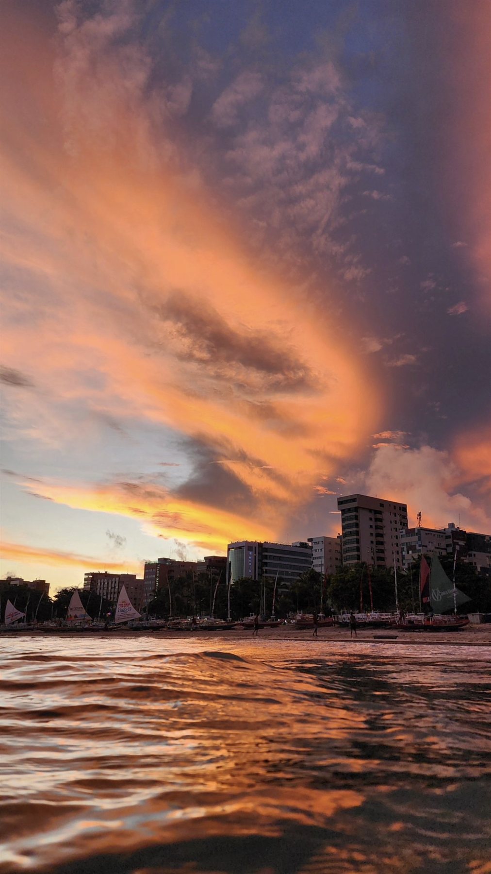 Pôr do sol dramático sobre a orla de Maceió com céu alaranjado e jangadas na praia