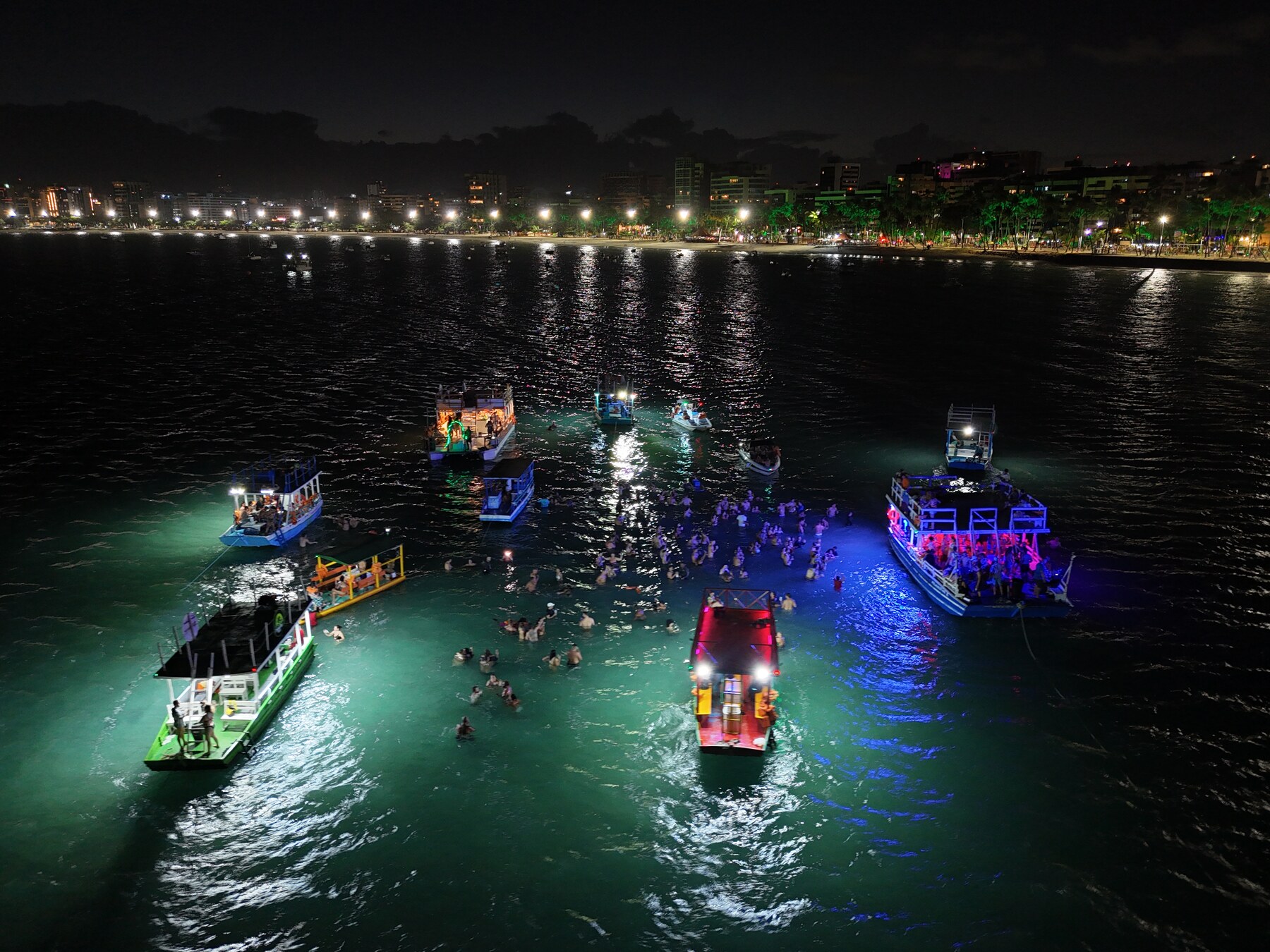 Vista aérea do banho de lua na Ponta Verde, Maceió, com barcos iluminados e pessoas na água